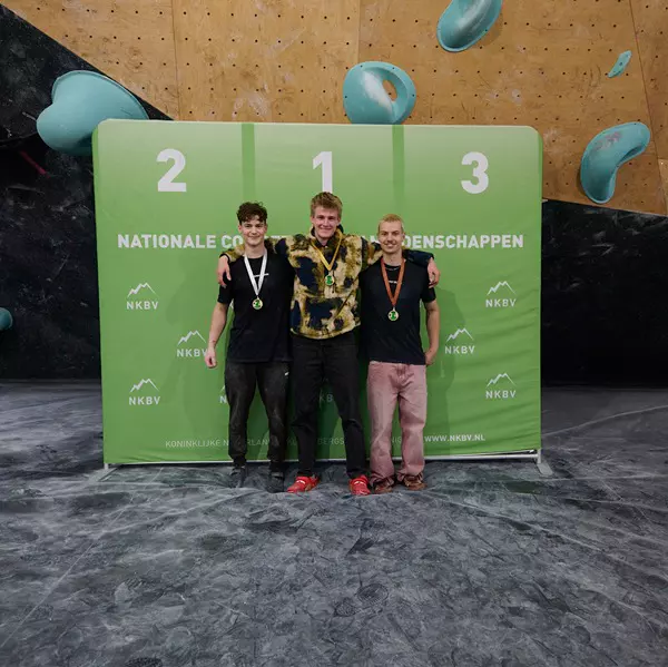 Drie jonge mannen met medailles staan op een podium voor een groen spandoek met de tekst ‘NKBV’ en ‘Nationale’, terwijl ze hun overwinning vieren tijdens een indoor klimwedstrijd. Achter hen zijn bouldergrepen aan de muur te zien.